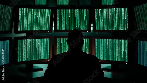 Silhouette of a person sitting at desk surrounded by multiple computer monitors displaying green digital data streams in dark room