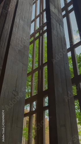 Weathered wooden beams in modern architectural installation. Sunlight streams through the gaps revealing green trees outside. 