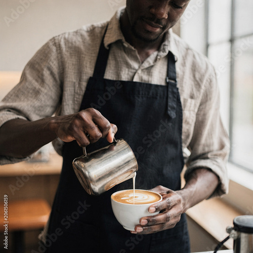 Professional African American barista pouring steamed milk into a white cup to create latte art in a modern coffee shop