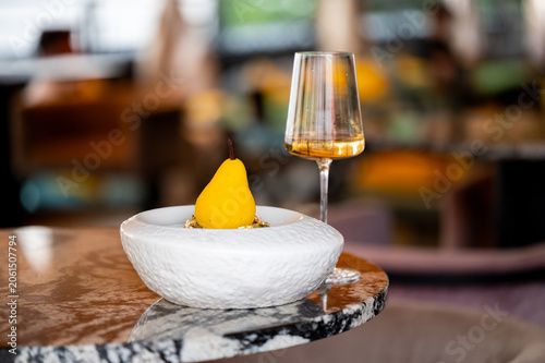 Konstfotografi A yellow pear-shaped dessert served in a white textured bowl next to a glass of white wine on a marble table