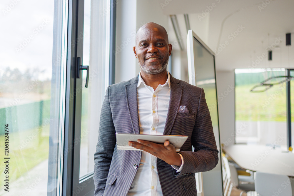 Fototapeta premium Mature black businessman smiling, holding tablet in modern office, portraying success and leadership