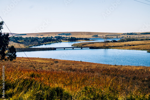 Fotografie Tranquil river landscape with a concrete bridge crossing a wide blue river surrounded by golden winter grass and rolling hills under a clear blue sky, eMalahleni, South Africa