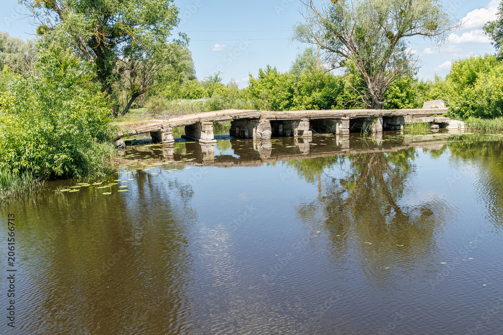 Obraz premium Old concrete low bridge crossing a calm river surrounded by green trees on a sunny summer day