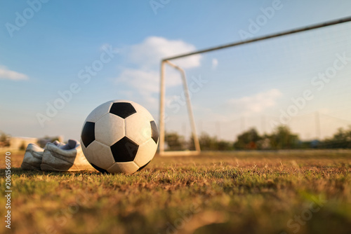 Football training and active lifestyle concept. Close up of a classic soccer ball and football boots on green grass field with blurred goal net in the background at sunset.