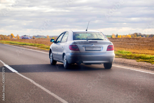 Silver sedan car driving away on rural asphalt road through countryside under cloudy sky, fields with dry grass and autumn trees along road