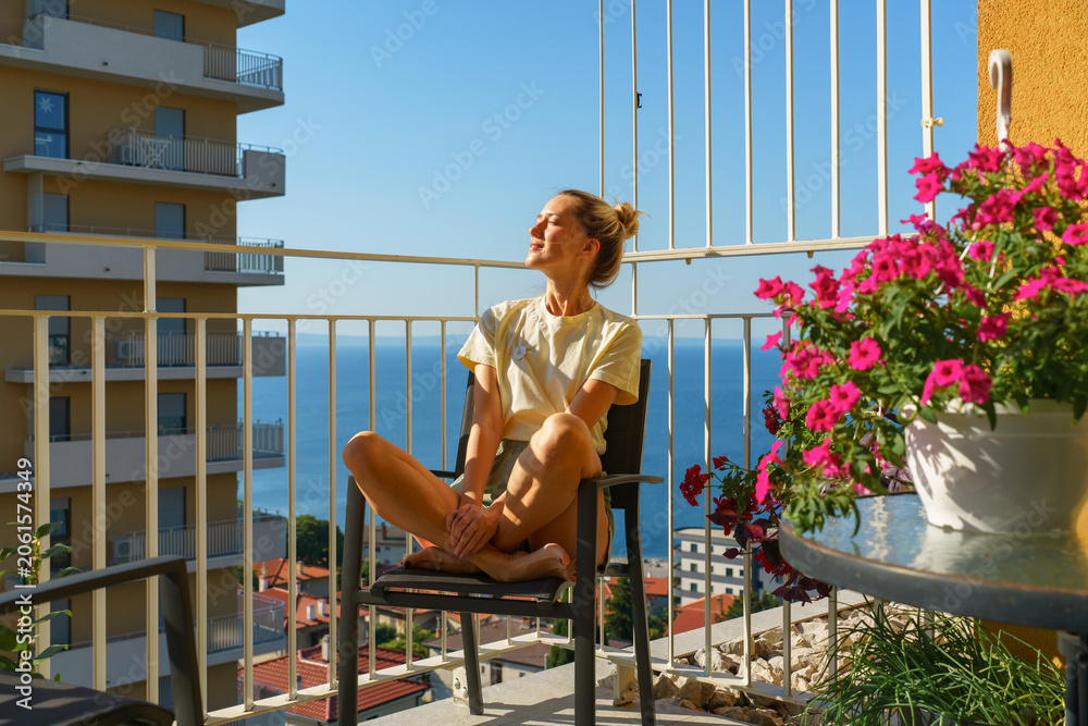 Obraz premium Woman relaxing on a sunny balcony with sea view and blooming flowers, enjoying mindfulness and wellness at home. Concept of slow living, self care, work life balance and summer lifestyle