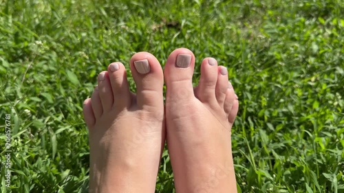 Close-up female feet with nude pedicure relaxing on green grass in summer, horizontal video