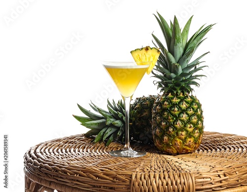 A refreshing cocktail in a martini glass with a pineapple wedge garnish, accompanied by two pineapples on a wicker surface