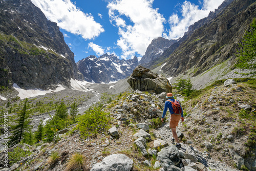 A hiker on a trail through the rugged glacial and rocky landscape of Écrins National Park in the French Alps. Large boulders, alpine vegetation, and scattered larch trees.