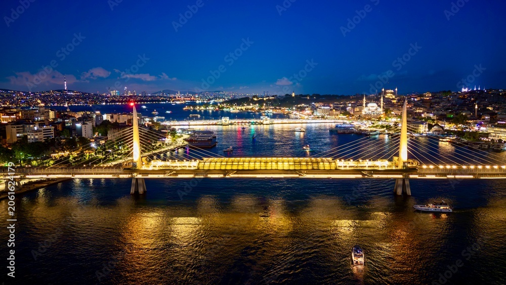 Fototapeta premium Istanbul, Turkey - June 13, 2026: Aerial drone night view of Haliç Bridge in Istanbul, Turkey, showing the bridge over the Golden Horn, city lights, waterfront districts, and reflections on the water.