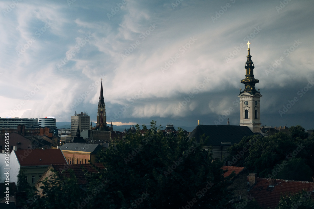 Obraz premium City skyline with church steeple and cloudy sky over rooftops, urban architecture under calm morning light glow.