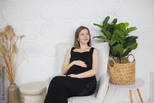 Pregnant woman sitting in cozy armchair, touch her belly in modern white interior. Thinking about future materhood.
