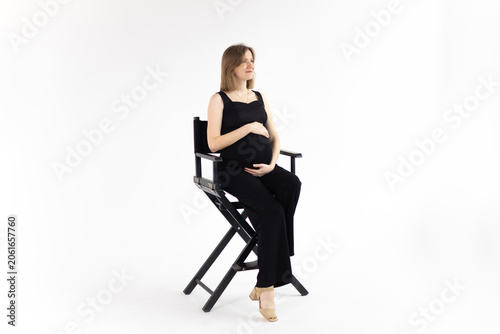 Smiling pregnant woman in black jumpsuit sitting on chair, holding her belly on white background.