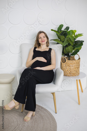 Happy pregnant woman sitting in an armchair and holding her belly, smiling at the camera.