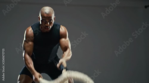 A powerful dark-skinned bald male athlete in a fitted tank top intensely performs rhythmic battle rope exercises against a dark gym background with artistic bokeh. Sweat glistens on his focused face,