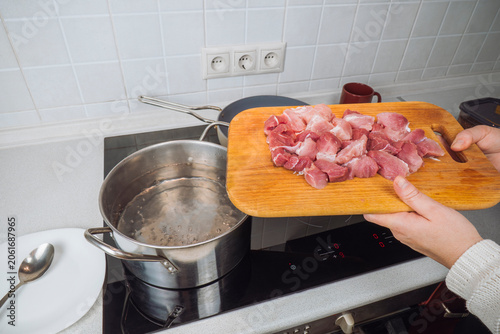 Woman preparing dinner by holding a wooden cutting board with chopped raw pork meat pieces over a stainless steel pot of boiling water on a stove