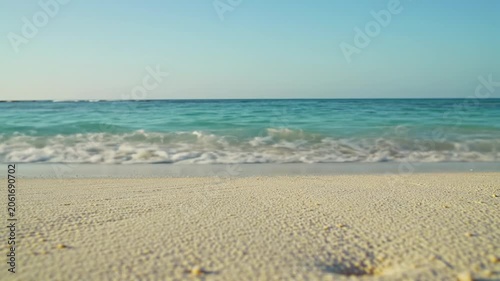 A wide establishing video of a pristine tropical beach with crystal-clear turquoise ocean waves and soft white foam washing onto untouched golden sand under a clear blue sky at golden hour, low-angle