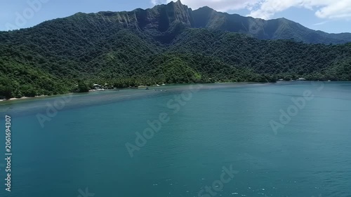 A cinematic drone video sweeps over a majestic tropical mountain range with lush green vegetation, descending to a calm azure bay. Low-angle tracking video glides along pristine water's edge,