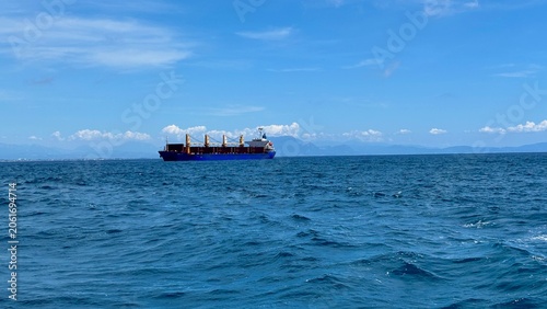 Fotografie Large cargo ship sailing on blue mediterranean sea under partly cloudy sky with