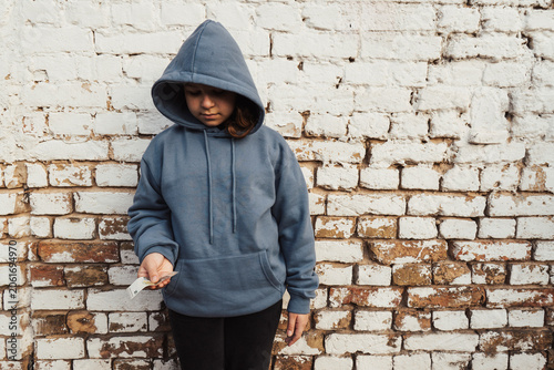 Teenage girl in a blue hooded sweatshirt holding a euro cash banknote while standing against a white weathered brick wall on the street