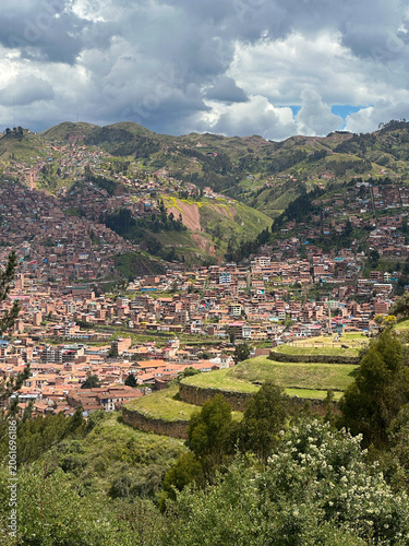 Panoramic view of Cusco, Peru, seen from the historic Sacsayhuamán archaeological site. The ancient Inca capital spreads across a mountain valley, surrounded by dramatic Andean landscapes and clouds.