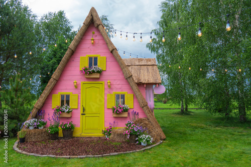Charming pink cottage decoration in a public park. A whimsical photo zone with a thatched roof, flowers, and string lights on a lawn under cloudy sky