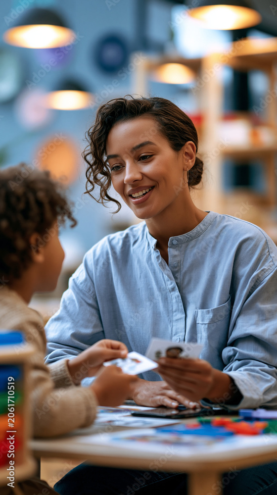 Fototapeta premium Wide shot of a child seated at a low table in a bright therapy room a speech therapist seated across from them holding picture cards and engaging the child in a structured