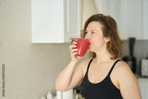 Fotografie Woman Drinking From Red Mug While Looking To Side In Bright Home Kitchen Interior Casual Sleeveless Top Short Brown Hair Domestic Morning Routine Standing By Countertop