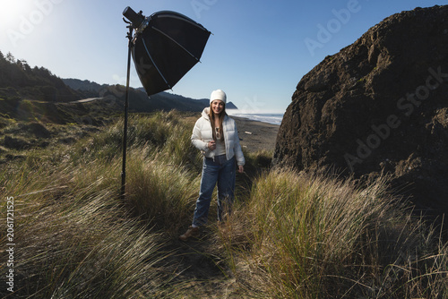 Woman stands on a sand dune in Gold Beach Oregon, with a strobe and softbox for a photoshoot.