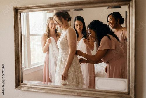 Bridesmaids helping the bride into her lace gown, seen in an antique mirror's reflection.