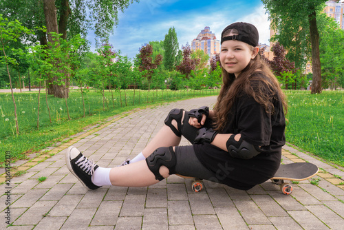 Smiling teenage girl with long hair wearing a black t-shirt, cap, and safety pads sits on a skateboard on a paved path in a green park on a cloudy day