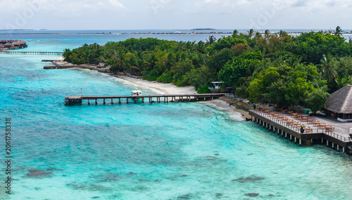 Photos Aerial view of a Maldives luxury resort with turquoise shallows, overwater piers, long wooden jetties, water villas, coral reefs, and a beachfront terrace under midday light