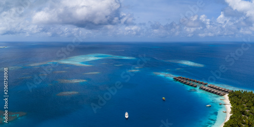 Bild auf Leinwand Aerial view of a Maldives luxury resort, overwater villas extend from a palm island into a turquoise lagoon, with sandbars, coral patches, boats, and a yacht visible