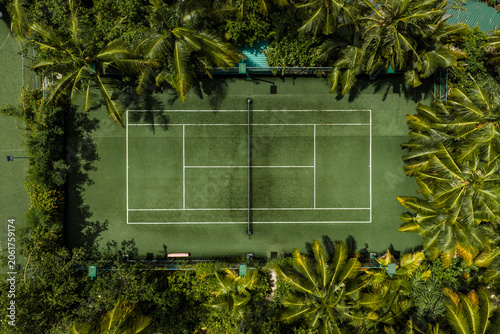 Aerial view of a secluded tennis court in the Maldives, white lines and net visible, surrounded by palm trees and foliage, bright daylight and even overhead lighting.