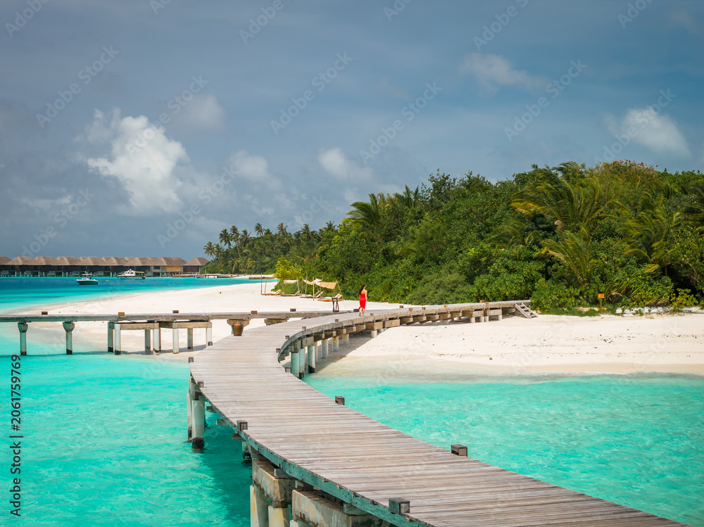 Fototapeta premium A curved wooden boardwalk arcs over shallow turquoise water toward a palm island. A single person walks along it. Overwater bungalows sit on the horizon at midday.