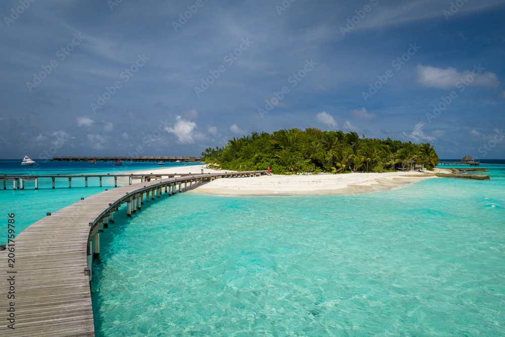 Fototapeta premium Sunlit Maldives resort shows S shaped wooden boardwalk over turquoise shallows toward white sand and palms, with overwater bungalows and a small boat under midday light.