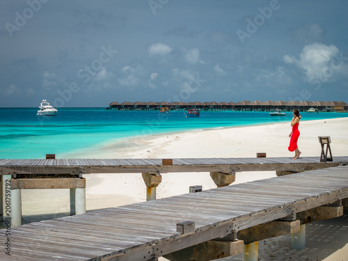 Cuadro en lienzo A woman walks along white sand by clear water and rustic boardwalks in the Maldives