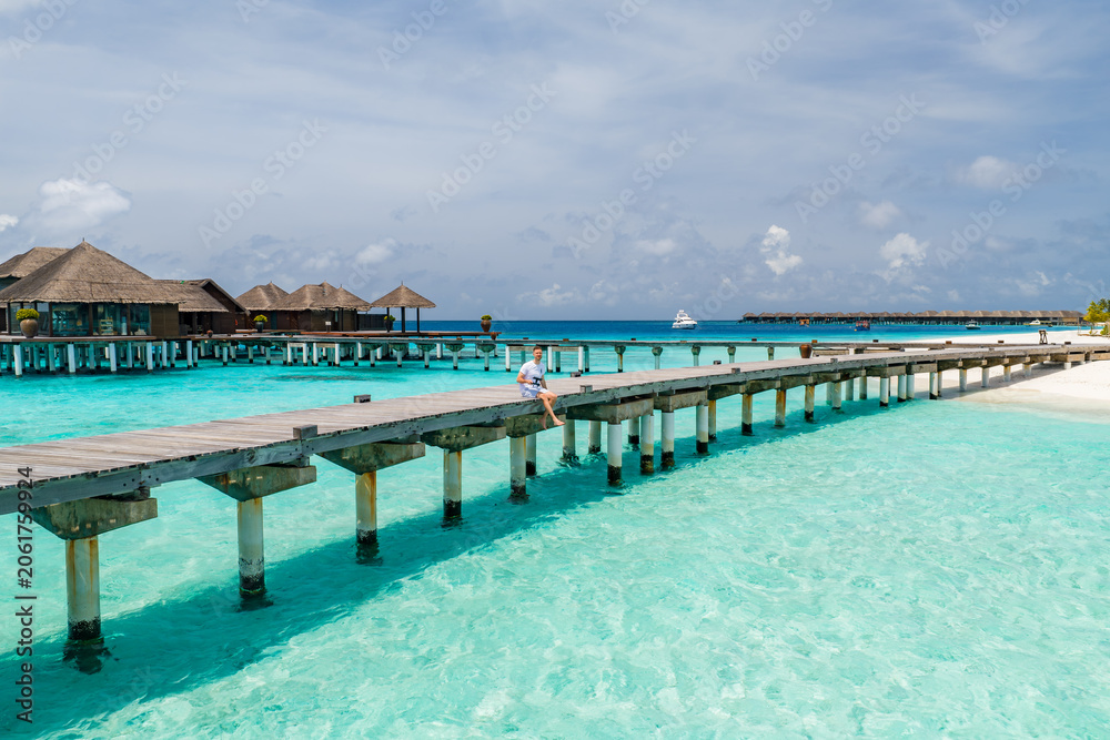 Fototapeta premium Sunlit wooden boardwalk crosses turquoise lagoon to thatched bungalows in Maldives. A person sits mid bridge, a white yacht glides on horizon, sandy beach lines right.