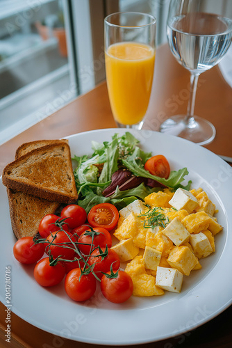 Breakfast plate featuring scrambled eggs, cherry tomatoes, salad, toast, and beverages including orange juice and water in a bright indoor setting