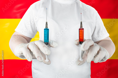 Medical worker in white t-shirt and gloves holding two syringes with red and blue vaccine or medicine on background of Spanish national flag