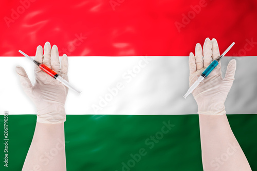 Hands in protective medical gloves holding syringes with red and blue liquid against the background of the national flag of Hungary