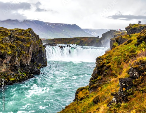 A serene waterfall cascades into a vibrant turquoise river surrounded by mossy cliffs and rugged mountains under a cloudy sky