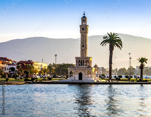 A serene waterfront scene featuring a tall, ornate tower with a clock face, surrounded by palm trees and buildings, set against a mountainous backdrop