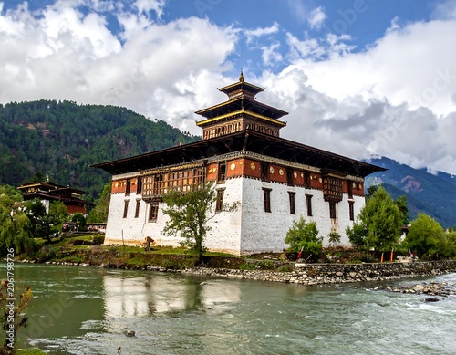 A serene white and brown building with a pagoda-style roof sits on the banks of a river, surrounded by lush green trees and majestic mountains under a blue sky with fluffy white clouds