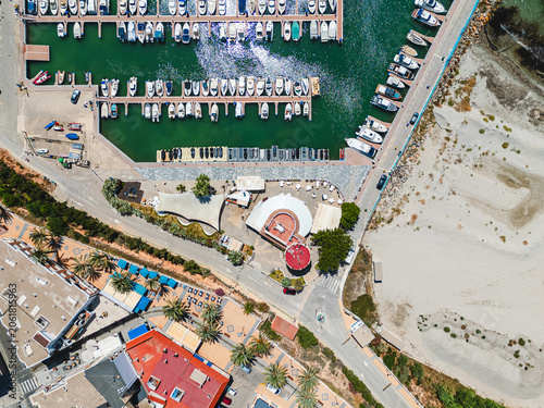 Aerial drone view of coastal marina with rows of moored boats in turquoise water, sandy beach with palm trees, waterfront buildings, seaside resort town of Torre de la Horadada. Costa Blanca. Spain