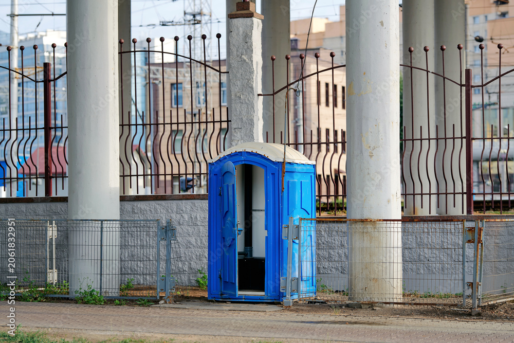 Naklejka premium Blue plastic portable toilet cabin with an open door. Public mobile restroom facility placed on city street pavement beneath massive overpass. Temporary sanitary water closet with an unlatched door