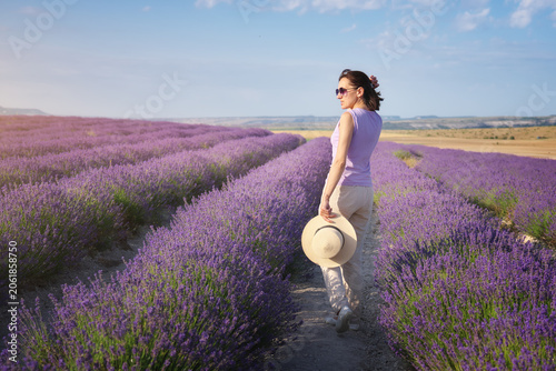 Young woman walking through rows of blooming violet lavender field holding a straw hat. Beautiful female tourist enjoying summer vacation