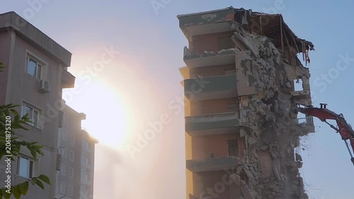 Demolition of building: excavator is tearing down the old typical Turkish multi-storey apartment, residential building in Istanbul, Turkey: slow motion. Setting sun is shining through dust