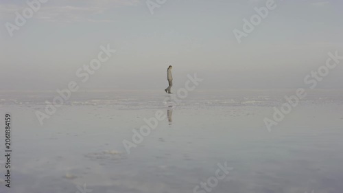 Individual in light clothing walks steadily across a reflective salt flat, with a vast, hazy sky creating a serene and tranquil atmosphere in the background