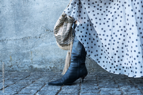 Fotografie Lower section shot focusing on a woman's black leather high-heel boot and the hem of a white chiffon skirt with black polka dots against a concrete background